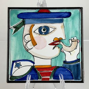 May include: Square ceramic tile depicting a stylized sailor's face with a blue hat, red and blue striped shirt, and a pipe. The artwork has a black border and a turquoise background. The sailor's face is painted with bold colours and simple lines.