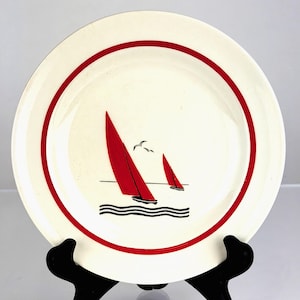 Puede incluir: Un plato de cerámica blanco con borde rojo y diseño náutico. El plato presenta dos veleros rojos sobre un mar azul, con dos gaviotas en el cielo. El plato se exhibe sobre un soporte negro.