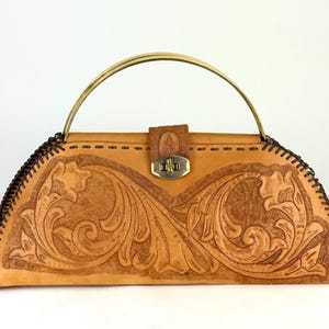 May include: Tan leather handbag with a brass handle and clasp. The bag features an embossed floral design on the front and is stitched with dark brown thread around the edges. The handle is arched and attached to the top of the bag.