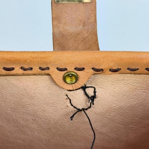 May include: Close-up of a tan leather bag with a brass buckle and snap closure. The bag features hand-stitched details and a visible tear near the snap. The leather has a natural, textured appearance.