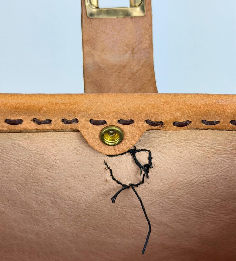 May include: Close-up of a tan leather bag with a brass buckle and snap closure. The bag features hand-stitched details and a visible tear near the snap. The leather has a natural, textured appearance.
