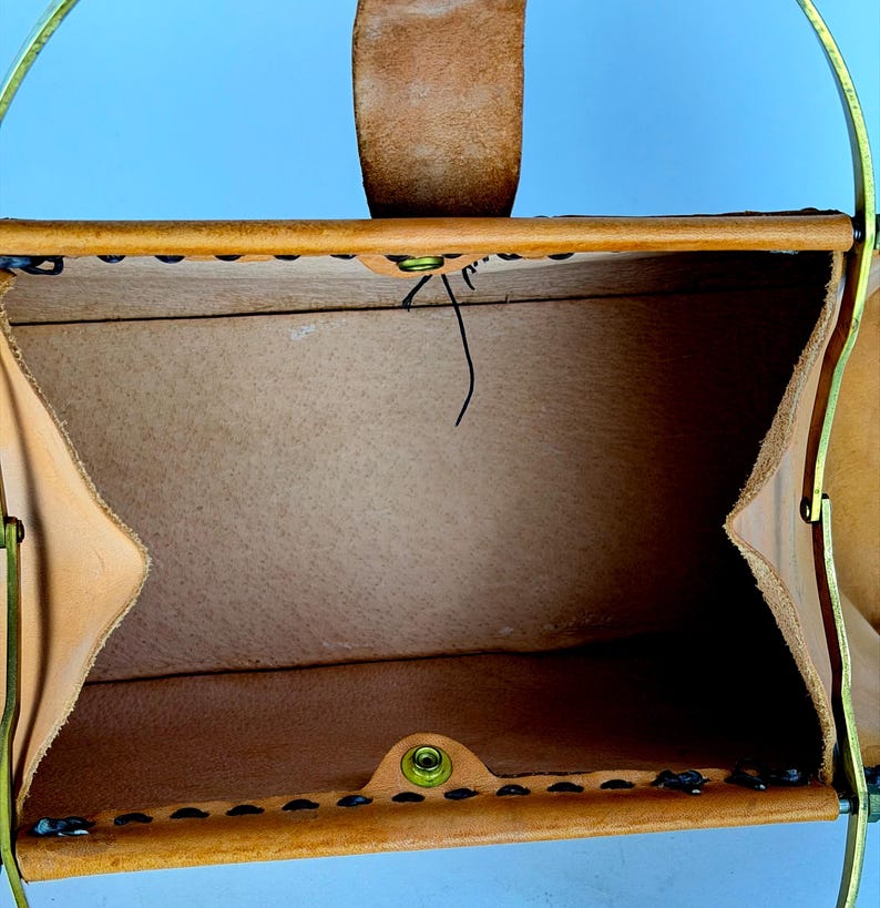 May include: Open view of a tan leather handbag with a brass handle and a leather strap. The bag is stitched with black thread and has a brass clasp. The interior is a lighter tan color.