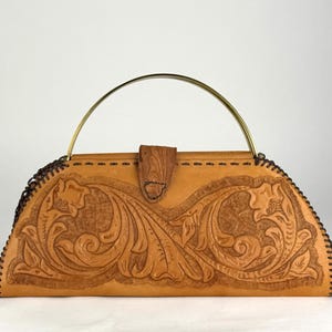 May include: A tan leather handbag with a floral embossed design. The bag has a curved shape, a brass handle, and a wooden clasp. The edges are stitched with dark brown thread. The bag is a vintage style.