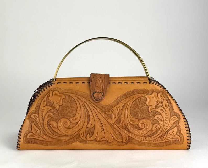 May include: A tan leather handbag with a floral embossed design. The bag has a curved shape, a brass handle, and a wooden clasp. The edges are stitched with dark brown thread. The bag is a vintage style.