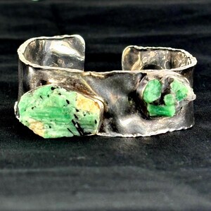 May include: A silver cuff bracelet featuring two raw emerald stones. The bracelet has a hammered texture and a wide band. The emeralds are a vivid green with black inclusions and a hint of yellow.