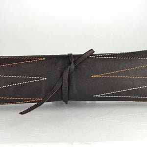 May include: A rolled-up, dark brown leather case with white and orange zigzag stitching. A matching leather tie wraps around the case. The case appears to be designed for storing art supplies or other small items.