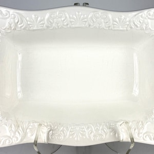 Puede incluir: Una fuente rectangular de color blanco roto con un borde decorativo en relieve con motivos florales y volutas. La fuente tiene bordes suavemente curvados y está sostenida por un soporte transparente. Adecuada para servir aperitivos o postres.