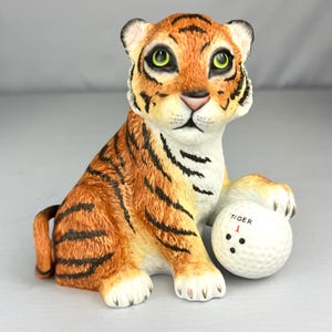 Puede incluir: Una figura de tigre de cerámica con pelaje naranja, rayas negras y ojos verdes, sentada en posición frontal. Una pata descansa sobre una pelota de golf blanca con la palabra TIGER en negro, un número 1 rojo y tres puntos negros.
