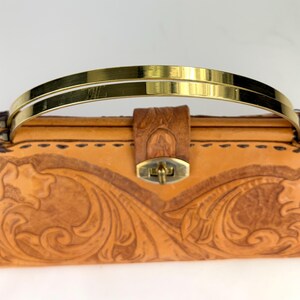 May include: A tan leather handbag with a gold-colored handle. The bag features intricate floral carvings and a brass clasp. The handle is a double bar design. The bag is a classic style.