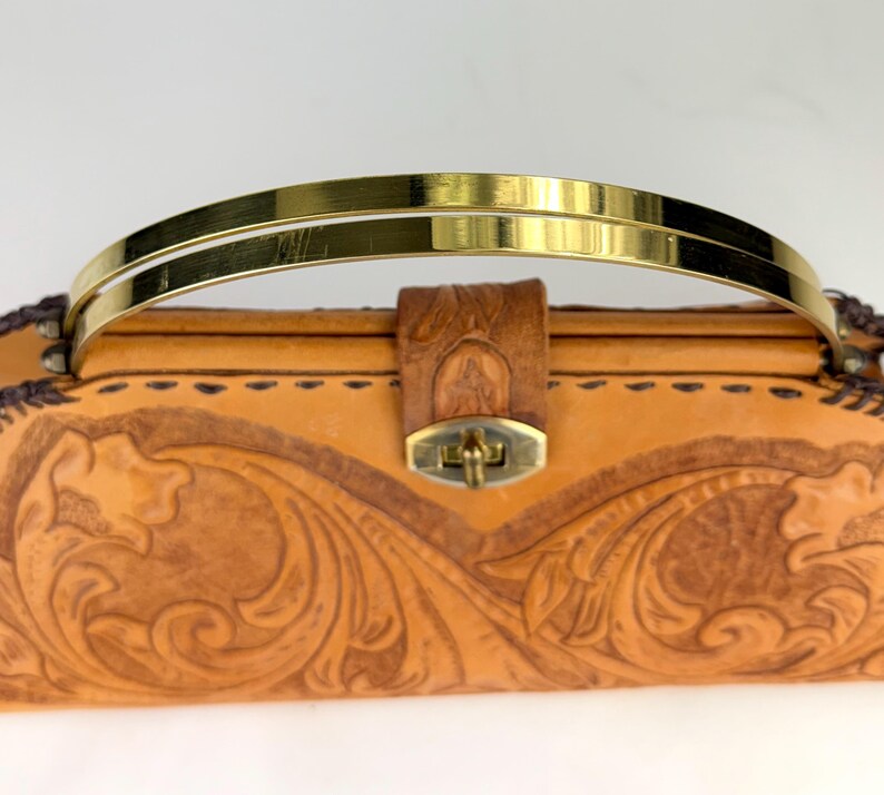 May include: A tan leather handbag with a gold-colored handle. The bag features intricate floral carvings and a brass clasp. The handle is a double bar design. The bag is a classic style.