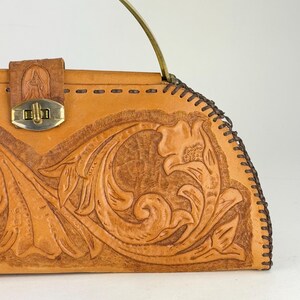 May include: A tan leather handbag with a floral embossed design. The bag features a brass clasp, a brass handle, and decorative stitching along the edges. The bag has a vintage aesthetic.