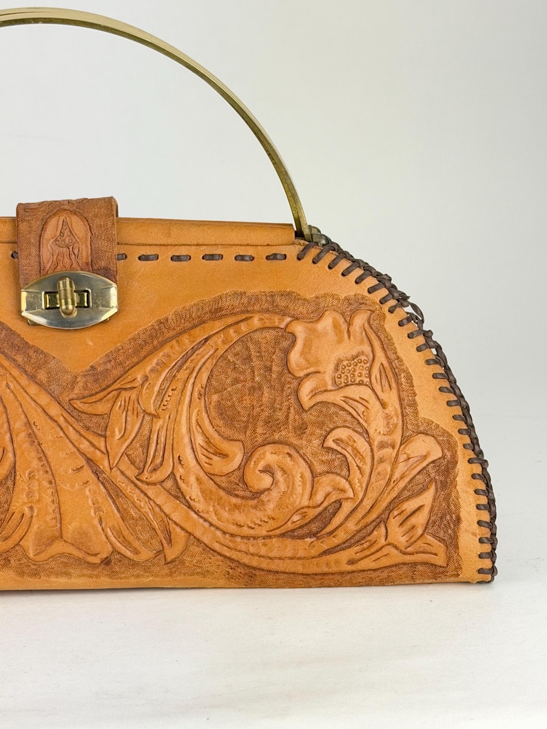 May include: A tan leather handbag with a floral embossed design. The bag features a brass clasp, a brass handle, and decorative stitching along the edges. The bag has a vintage aesthetic.
