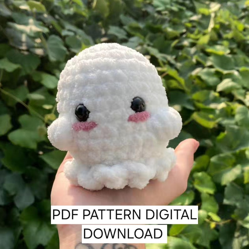 K&ouml;nnte beinhalten: Ein wei&szlig;es, geh&auml;keltes Geister-Amigurumi-Pl&uuml;schtier mit schwarzen Augen und rosa Wangen. Das Spielzeug wird in einer Hand gehalten. Der Text "PDF PATTERN DIGITAL DOWNLOAD" befindet sich am unteren Rand des Bildes.