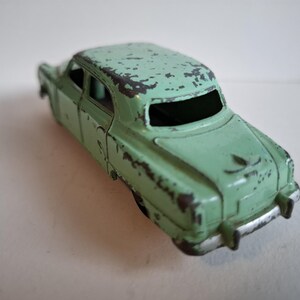 May include: A distressed, mint green toy car. The classic sedan shows signs of wear, with chipped paint revealing the metal beneath. The car has a black window and a rear bumper. The overall appearance suggests a vintage item, with a weathered aesthetic.