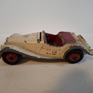 Op de afbeelding: Vintage speelgoedauto in crème en rood. De klassieke roadster heeft een crèmekleurige carrosserie met rood interieur en wielen. Het nummer "23" is zichtbaar op de zijkant. De speelgoedauto is ongeveer 13 cm lang.