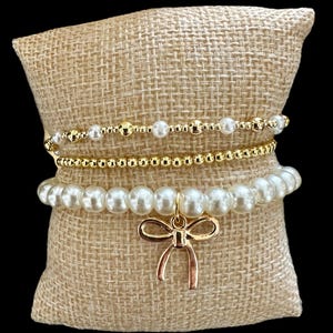 Puede incluir: Tres pulseras expuestas sobre un cojín de arpillera. La pulsera superior tiene pequeñas cuentas doradas y perlas blancas. La pulsera del medio está hecha de pequeñas cuentas doradas. La pulsera inferior tiene perlas blancas grandes y un dije de lazo dorado.