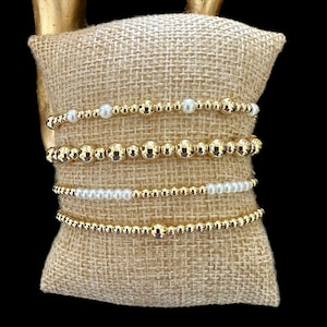 May include: Four gold bead bracelets stacked on a burlap cushion. The bracelets are made of small gold beads and some have white pearl beads.