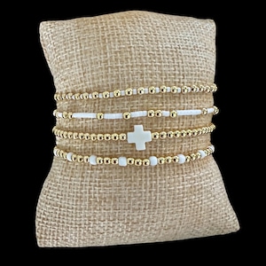 Op de afbeelding: Vier armbanden met kralen in goud en wit worden getoond op een jute kussen. De armbanden hebben gouden kralen, witte kralen en een witte kruisbedel. De armbanden zijn op elkaar gestapeld en tonen de verschillende ontwerpen.