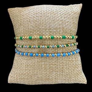 May include: Three beaded bracelets in shades of green and blue, displayed on a tan burlap pillow. The bracelets feature small, colorful beads interspersed with gold-colored beads. The bracelets are stacked on top of each other.