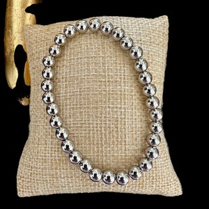 May include: A silver beaded bracelet with a shiny finish. The bracelet is made of round beads and is lying on a beige fabric surface.