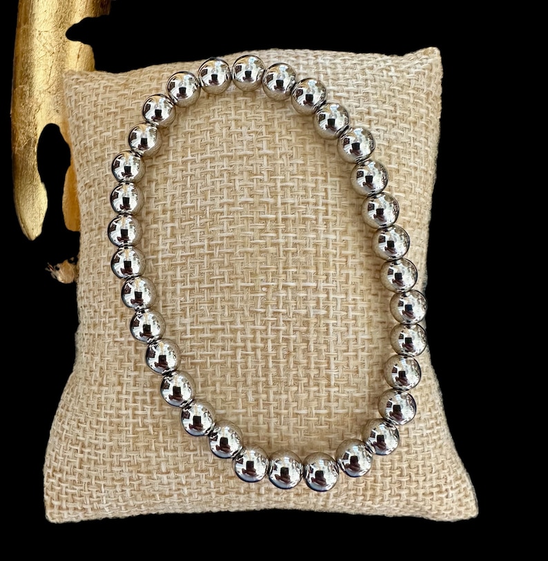 May include: A silver beaded bracelet with a shiny finish. The bracelet is made of round beads and is lying on a beige fabric surface.