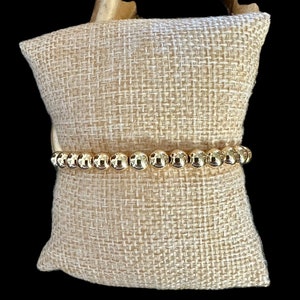 May include: A gold beaded bracelet on a beige fabric background.