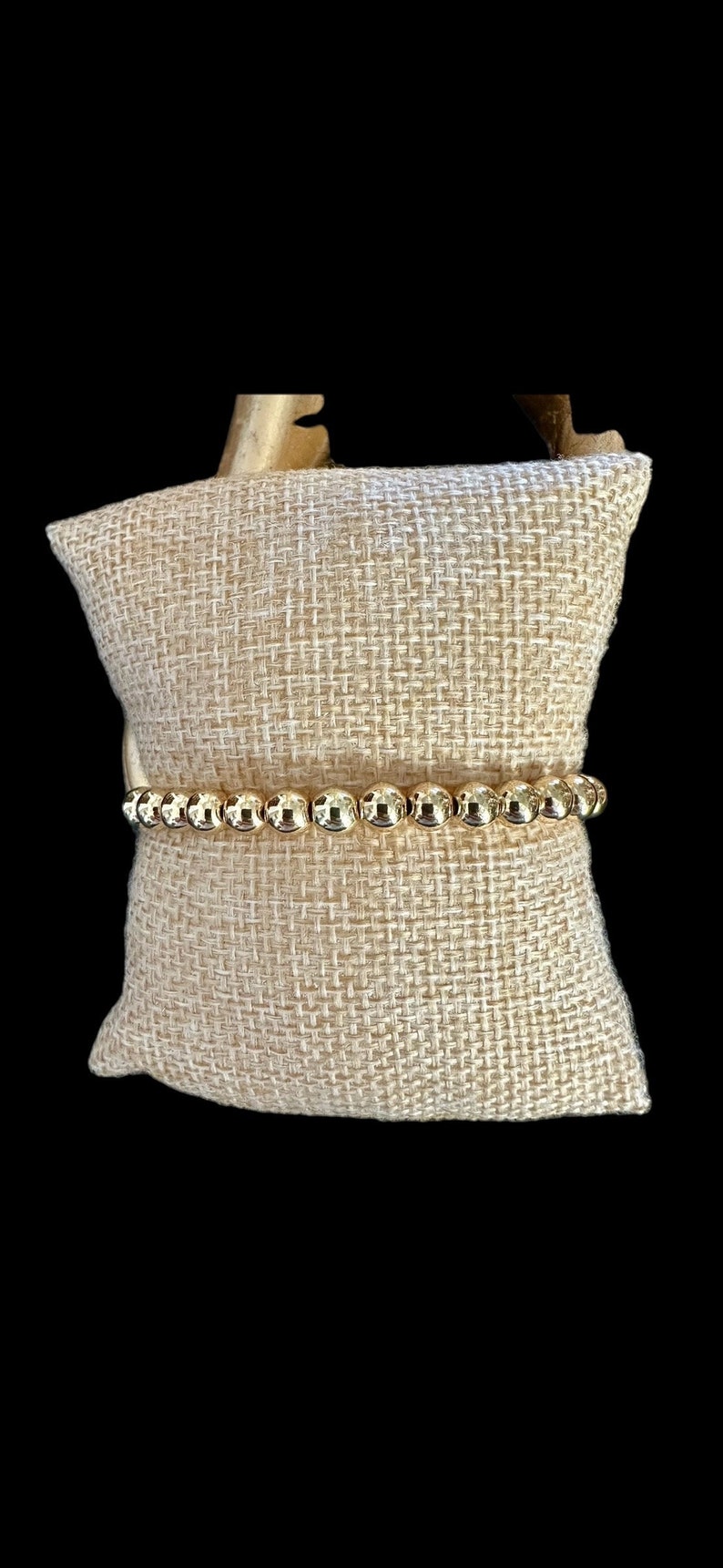 May include: A gold beaded bracelet on a beige fabric background.