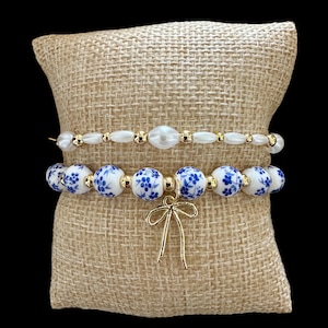 Puede incluir: Dos pulseras de cuentas. La pulsera superior tiene cuentas blancas y detalles dorados. La pulsera inferior tiene cuentas blancas y azules con estampado floral y un dije de lazo dorado. Las pulseras se exhiben sobre una almohada de arpillera.