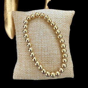May include: A gold beaded bracelet on a beige hessian cushion. The bracelet is made of round, shiny gold beads.