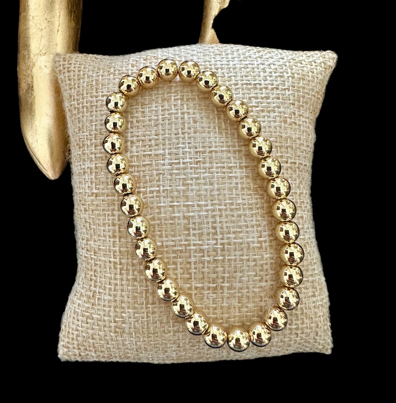 May include: A gold beaded bracelet on a beige hessian cushion. The bracelet is made of round, shiny gold beads.