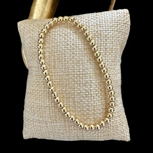 May include: A gold beaded bracelet on a beige fabric background.
