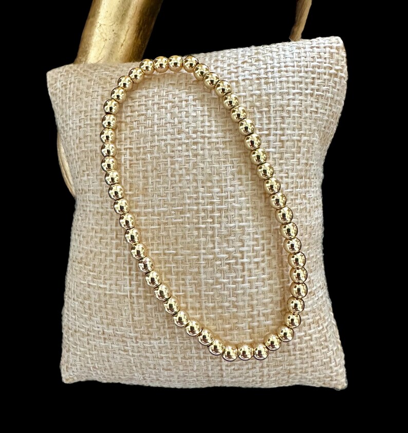 May include: A gold beaded bracelet on a beige fabric background.
