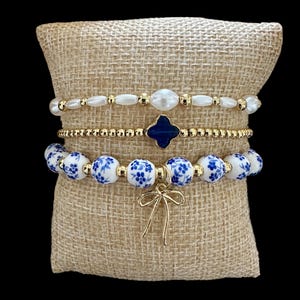 Puede incluir: Un conjunto de cuatro pulseras con un patrón floral azul y blanco, cuentas doradas y un dije de lazo dorado. Las pulseras están apiladas sobre una superficie de tela beige.