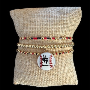 May include: A set of three bracelets. The bracelets have gold-coloured beads and red and black bead accents. The central charm is white with a baseball design, the number "#1" in black, and red stitching.