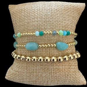May include: A set of three gold beaded bracelets with green and blue gemstone accents. The bracelets are stacked on a burlap cushion.
