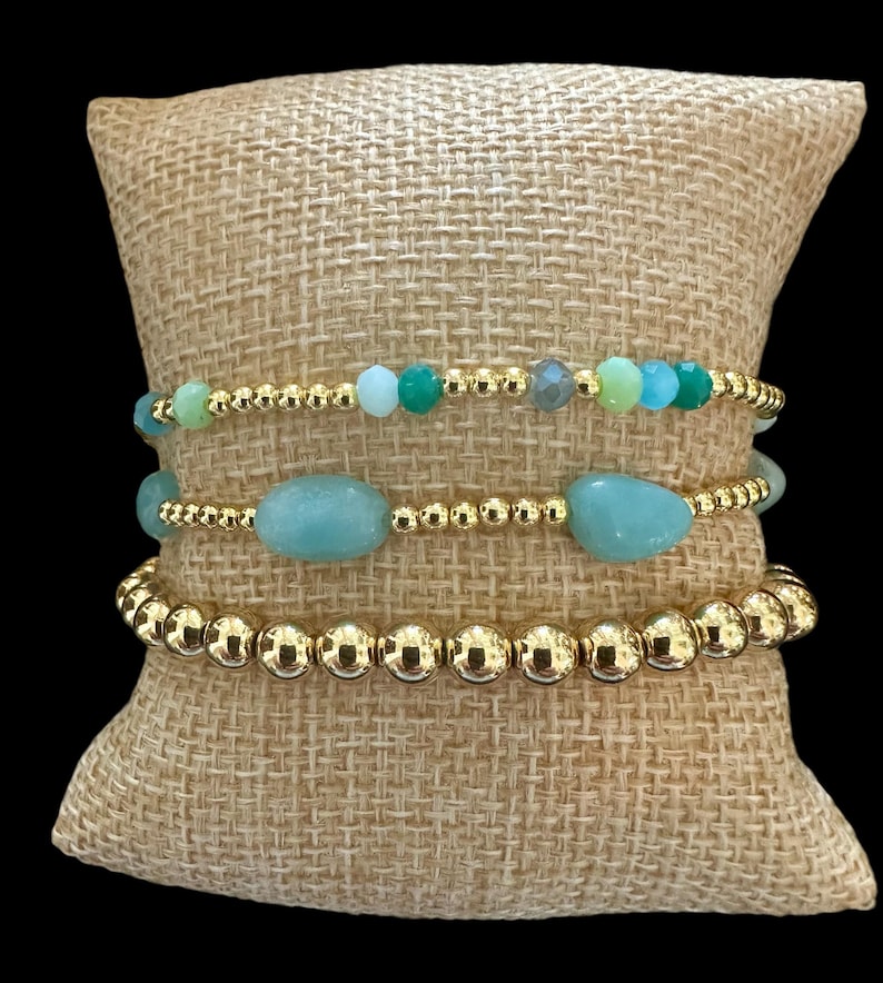 May include: A set of three gold beaded bracelets with green and blue gemstone accents. The bracelets are stacked on a burlap cushion.