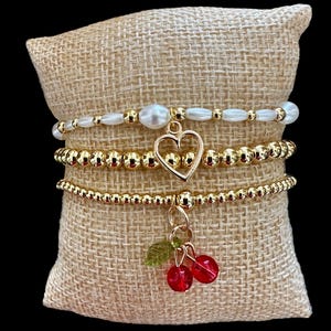 May include: Four gold bead bracelets with charms. The top bracelet is white with pearl-like beads. The second bracelet has a gold heart charm. The third bracelet has a cherry charm with red glass cherries and green leaves. The bracelets are displayed on a brown burlap surface.