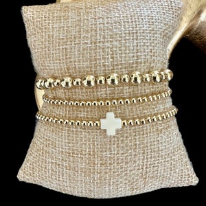May include: Three gold beaded bracelets stacked on a burlap surface. The top bracelet features a white cross charm.