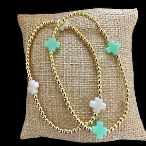 May include: Two gold beaded bracelets with cross-shaped charms in white and mint green. The bracelets are displayed on a tan, textured surface, creating a simple yet elegant presentation. The gold beads add a touch of sparkle.