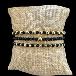 May include: Three beaded bracelets with gold and black beads are displayed on a burlap cushion against a black backdrop. The bracelets are stacked, showcasing a mix of gold and black beads in various sizes and designs. The bracelets are a stylish accessory.