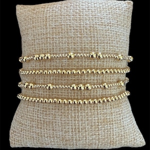 Può includere: Un set di quattro braccialetti di perline color oro con perline sferiche lucide di varie dimensioni disposte in motivi distinti, esposti orizzontalmente su un cuscino in tessuto beige chiaro testurizzato su uno sfondo nero.