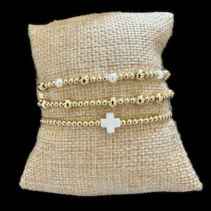 May include: Three gold beaded bracelets. One bracelet features small white pearls, another has a white cross charm. The bracelets are displayed on a beige burlap pillow against a black background.