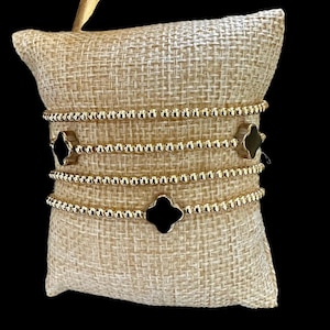 May include: Four gold beaded bracelets with black clover-shaped charms are displayed on a burlap pillow. The bracelets are made of small, round gold beads. Two bracelets have a black clover charm. The pillow is beige.