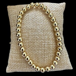 May include: A gold beaded bracelet with round beads. The bracelet is on a beige fabric surface.