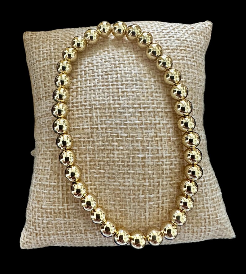 May include: A gold beaded bracelet with round beads. The bracelet is on a beige fabric surface.