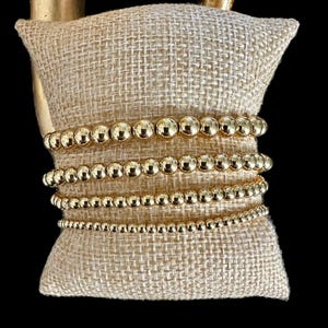 May include: A set of four gold beaded bracelets stacked on a beige fabric surface. The bracelets are made of small, round beads.