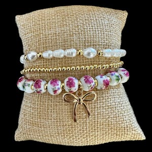 Puede incluir: Una pila de tres pulseras exhibidas sobre una almohada de arpillera. La pulsera superior presenta perlas blancas y cuentas doradas. La pulsera del medio está hecha de pequeñas cuentas doradas. La pulsera inferior tiene cuentas florales blancas y un dije de lazo dorado.