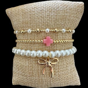 Puede incluir: Un conjunto de tres pulseras con cuentas doradas, perlas blancas y un dije de cruz rosa. Las pulseras se muestran en un cojín de arpillera con un dije de lazo dorado.