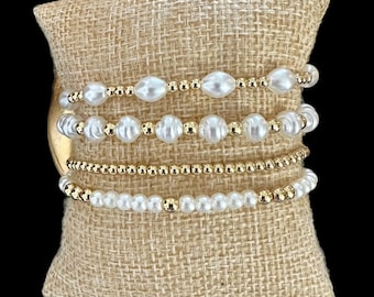 Può includere: Un set di cinque braccialetti con una combinazione di piccole perline dorate e perle finte bianche. I braccialetti sono impilati su un cuscino di juta marrone.
