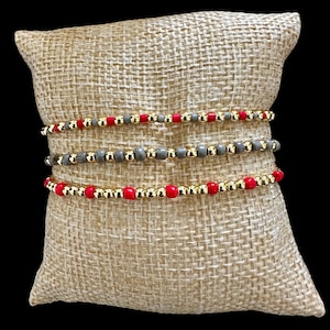 May include: Three beaded bracelets stacked on a burlap cushion. The bracelets are made of gold and red beads.